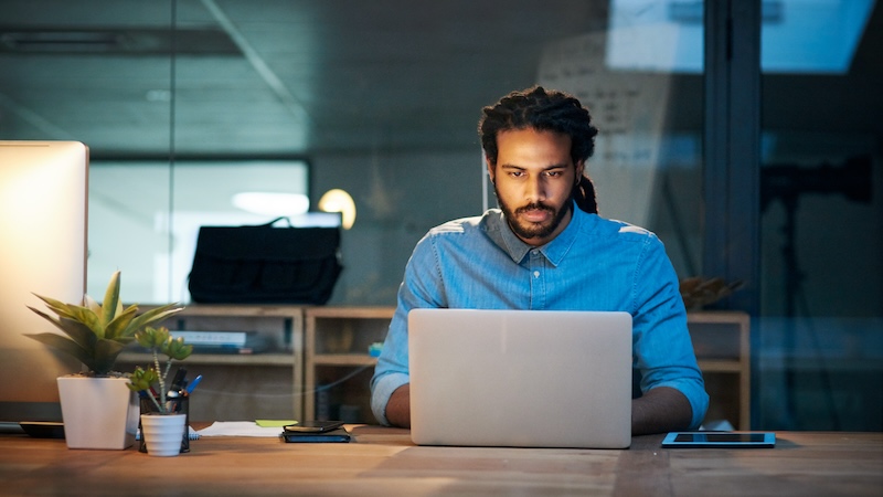 Man with dreadlocks wearing a denim shirt working on a laptop at a desk, surrounded by a smartphone, tablet, and plant, with a blurred background.