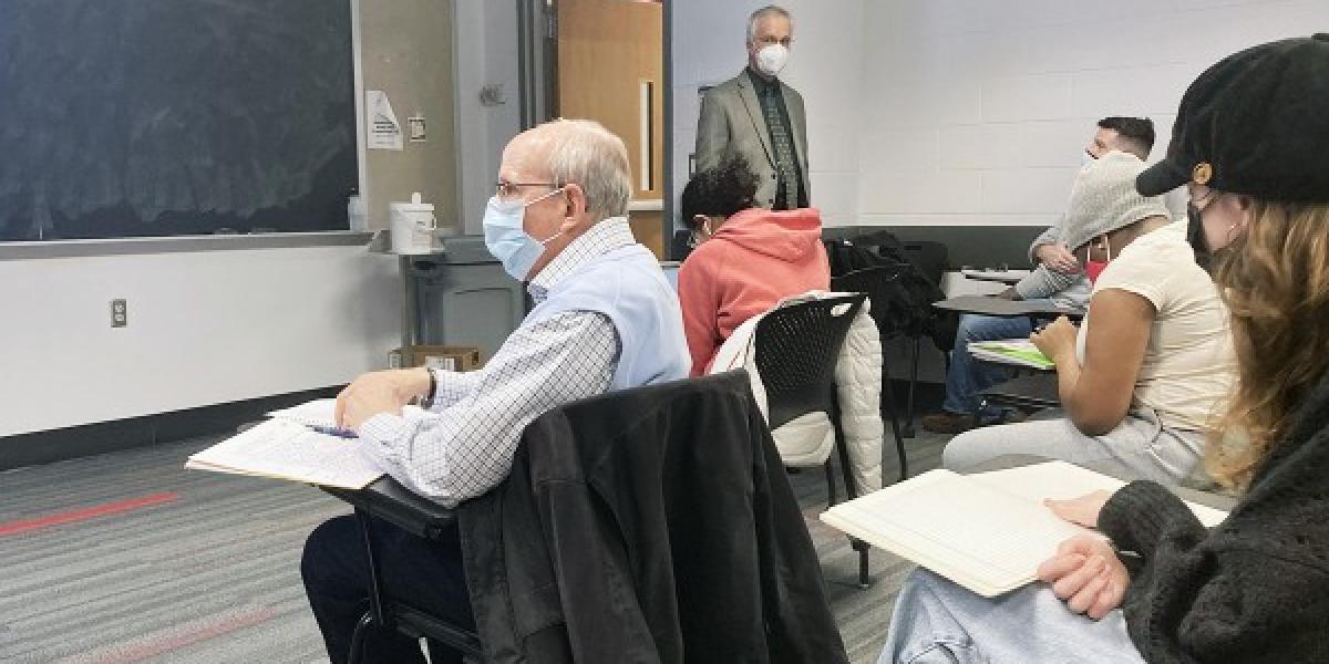 An older man with white hair sits in a desk at the front of a college classroom, wearing a surgical mask.